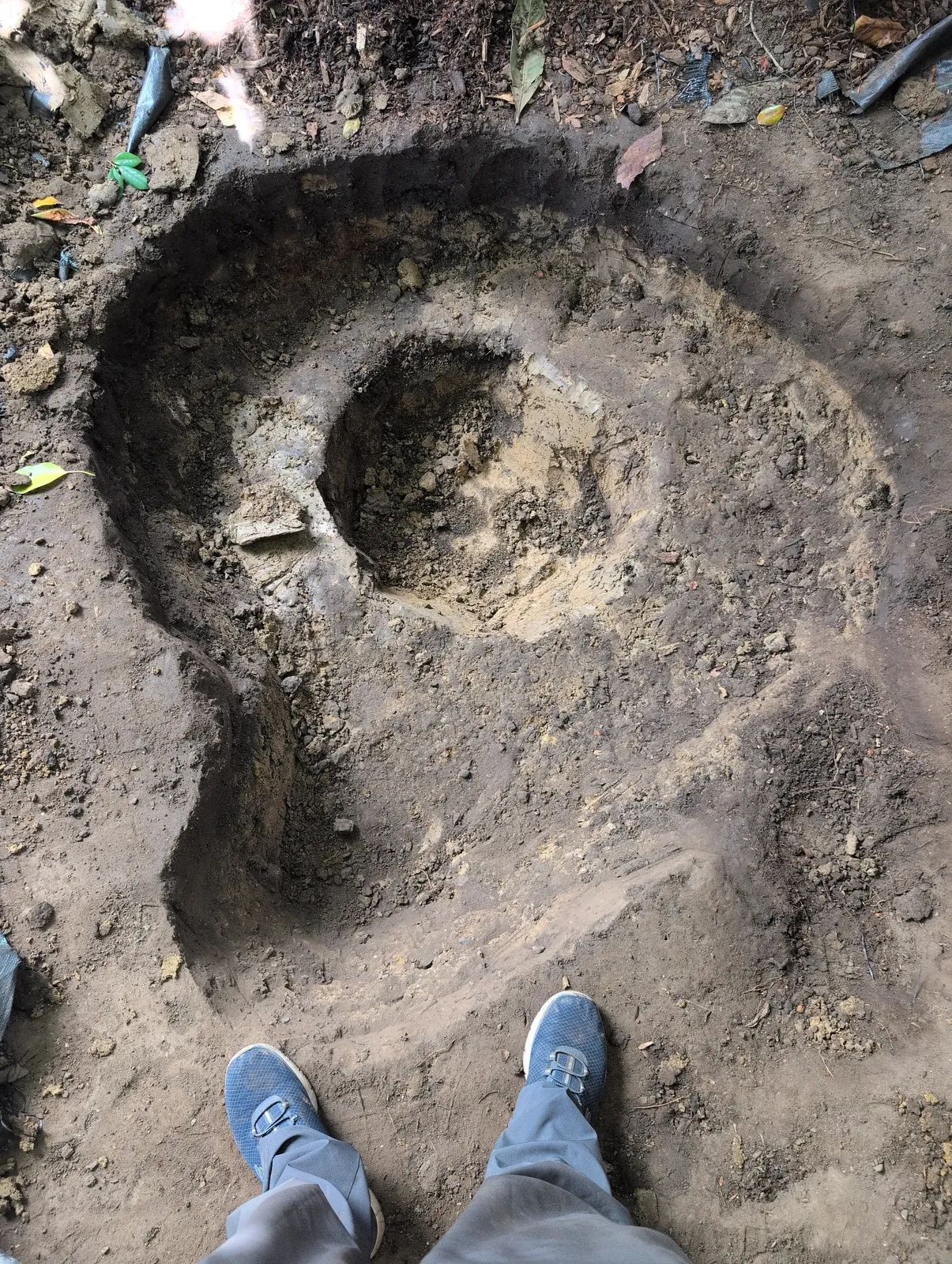 Looking down into the partly dug pond from above, showing the wide outer ledge and a deeper central bowl, with the builder's shoes at the bottom edge of the frame.