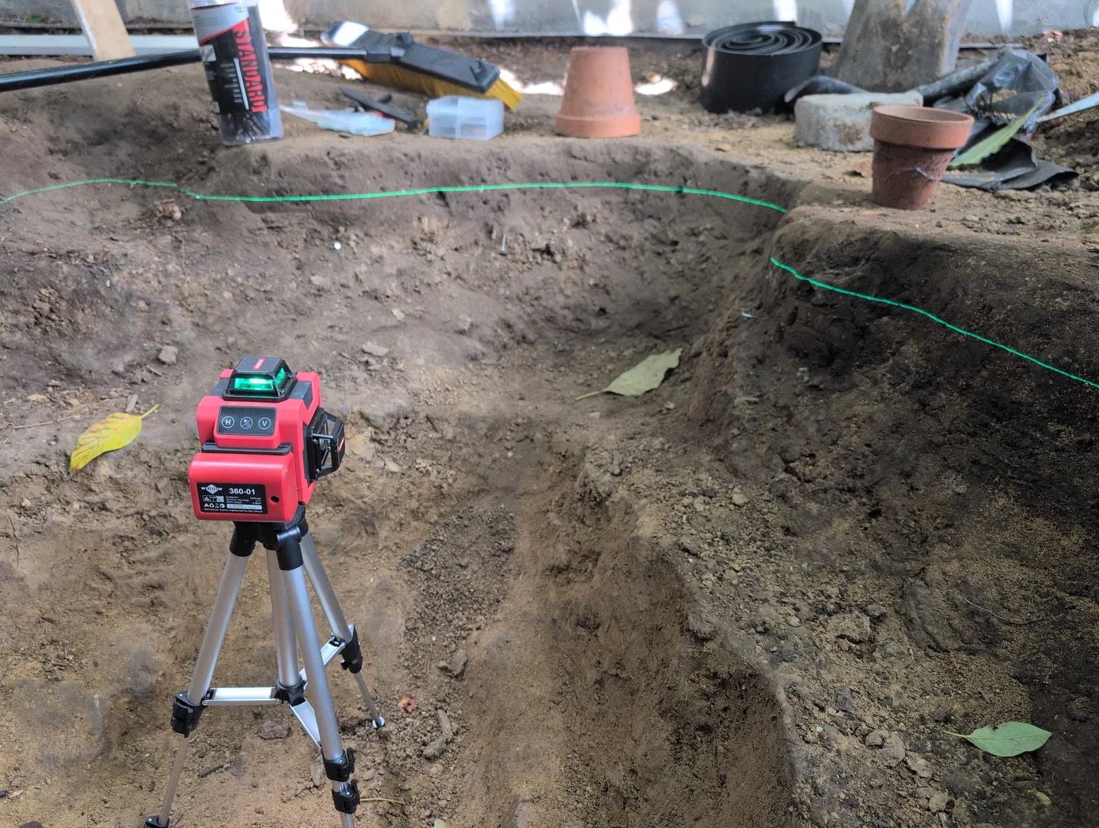 A red rotary laser level on a tripod beside the excavation, throwing a bright green horizontal line across the clay walls of the hole to mark the intended waterline.