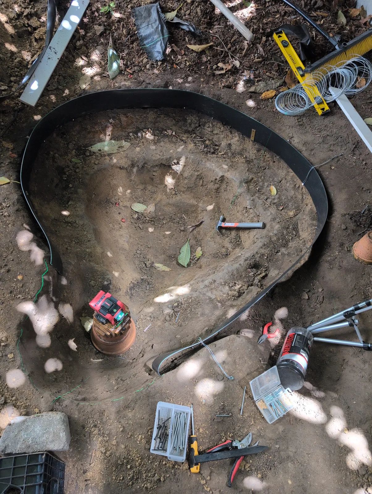 Top-down view of the excavation with a continuous black poly edging strip pinned around the rim in a curved trilobe outline, tools and wire staples scattered on the ground around it.