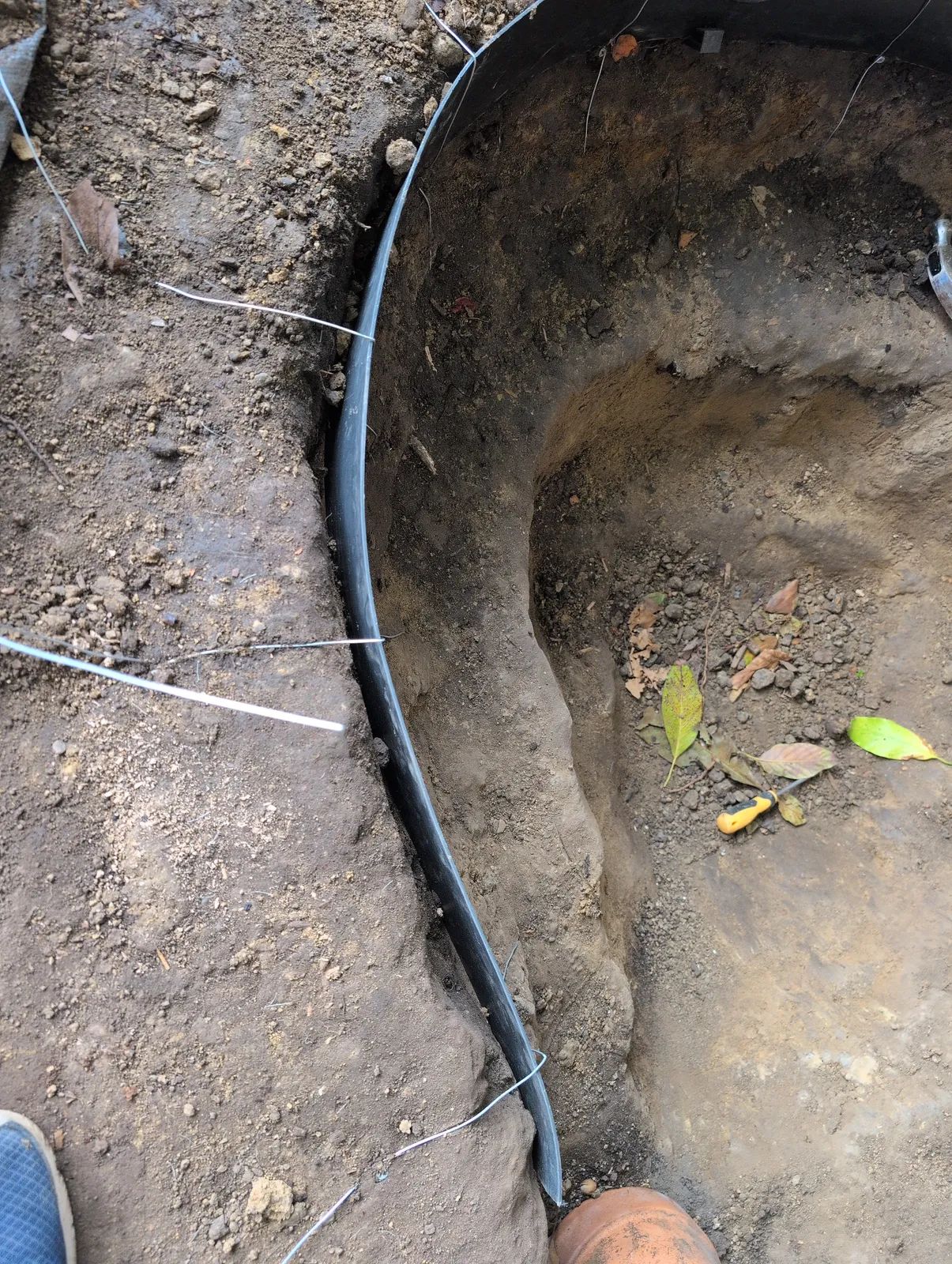 Close view of the black poly edging pinned into the clay rim with wire staples, showing a narrow crumbly gap between the edging and the cut clay wall behind it.