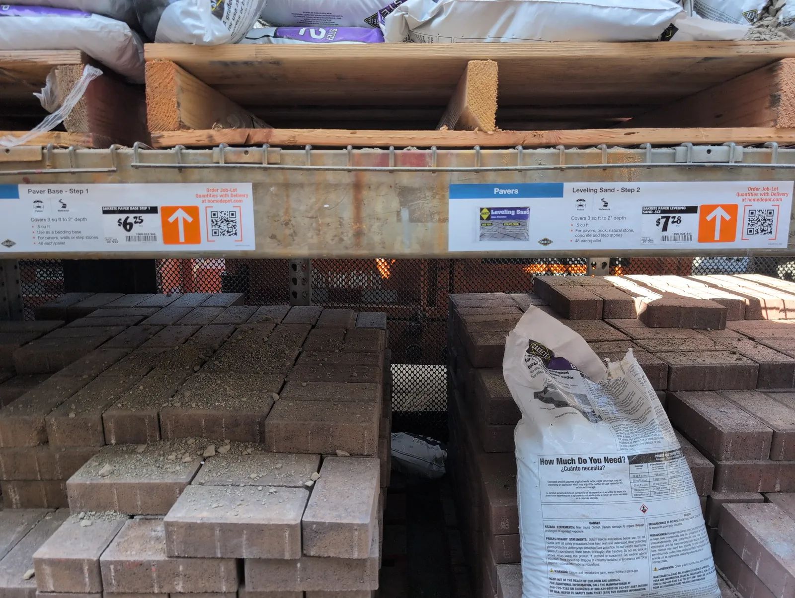 A hardware store rack with shelf labels reading Paver Base Step 1 at $6.25 and Leveling Sand Step 2 at $7.28, stacked pavers below and bagged material above.