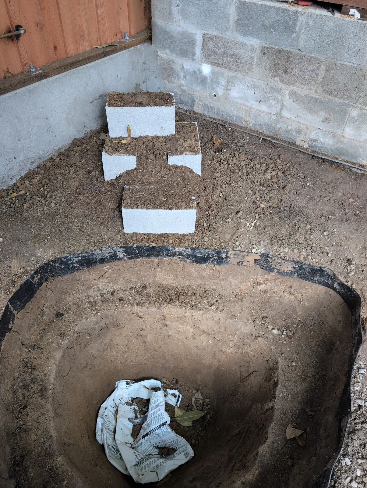 Three cinder blocks set in a stepped arrangement in the corner behind the excavated pond, forming the structural core of the waterfall, with the black poly edging ring visible around the rim below.