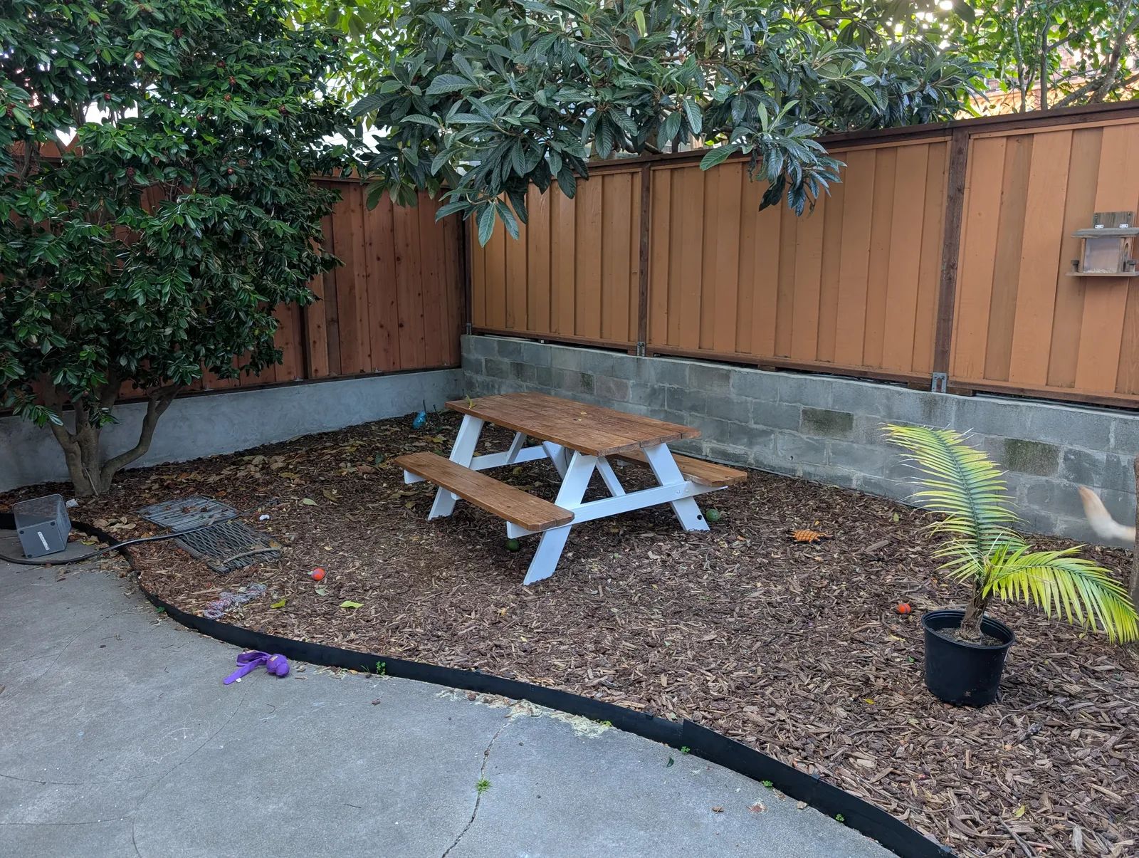 The untouched corner before any work: a picnic table on bark mulch, cinder block retaining wall along the back, wooden fence above, loquat branches overhanging from the left.