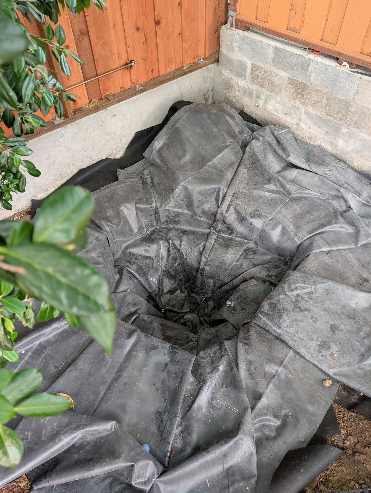 Black EPDM liner draped loosely into the excavation with large soft folds, spilling out over the surrounding ground and up against the cinder block and concrete retaining walls, before any water has gone in.